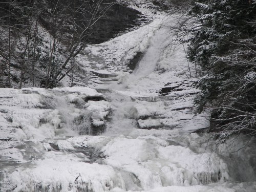 A frozen waterfall cascading over rocky terrain, surrounded by snow-covered trees and a winter landscape.