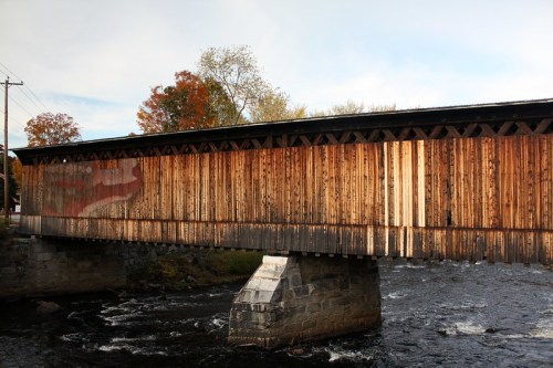 A wooden covered bridge spans a river, surrounded by autumn foliage and a clear sky.