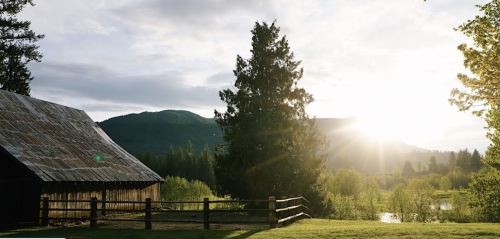 A rustic barn beside a lush green field, with mountains and a sunset in the background.