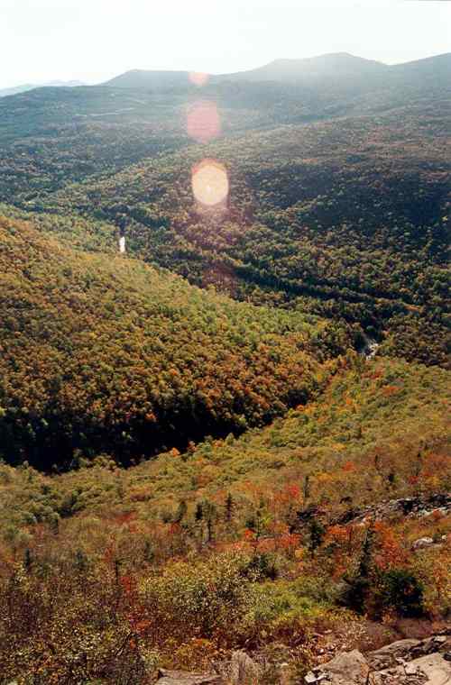 A panoramic view of a lush, green valley with autumn foliage and distant mountains under a bright sky.