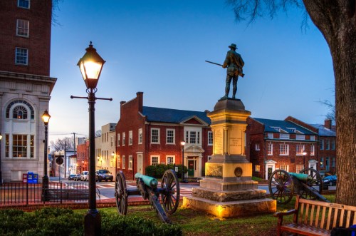 A statue of a soldier stands on a pedestal, surrounded by cannons and historic buildings at dusk.
