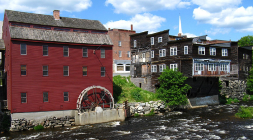 Historic red mill with a water wheel beside a river, surrounded by wooden buildings and greenery under a blue sky.