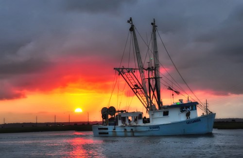 A fishing boat named Cassandra sails on water at sunset, with vibrant orange and purple clouds in the sky.