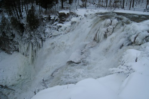 A frozen waterfall surrounded by snow-covered trees, with icy formations and rushing water cascading down.