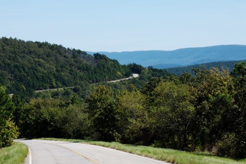 A winding road through lush green hills, with distant mountains under a clear blue sky.