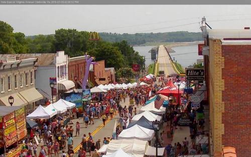 A bustling street fair with numerous tents, vendors, and crowds, set against a backdrop of trees and a distant road.