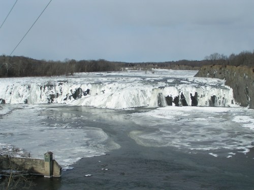 A frozen waterfall cascading over rocky cliffs, surrounded by icy terrain and a river below, under a cloudy sky.