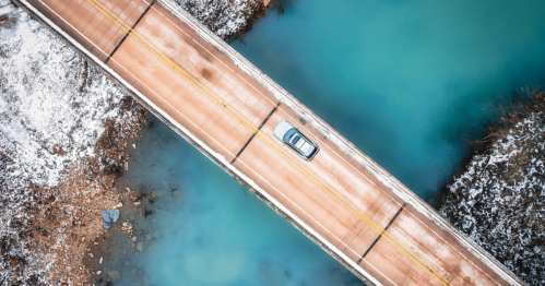 Aerial view of a car driving on a snowy bridge over a turquoise river.