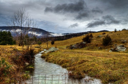 A serene landscape featuring a winding stream, grassy fields, and distant mountains under a cloudy sky.