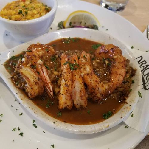 A plate of shrimp in a savory sauce, garnished with herbs, served alongside a bowl of corn and a lemon wedge.