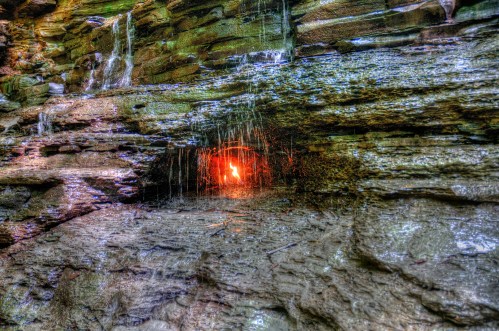 A rocky cliff with water trickling down, revealing a glowing orange light in a crevice.