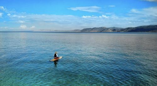 A person sits on a small boat in calm waters, surrounded by mountains and a clear blue sky.