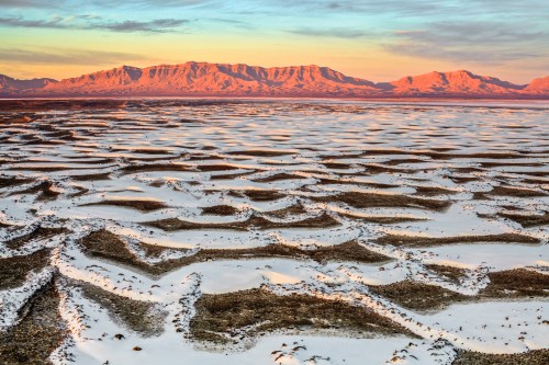 A vast, textured landscape of snow and rock formations under a colorful sunset with distant mountains.