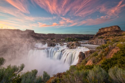 A stunning waterfall cascades over rocks, surrounded by lush greenery and a colorful sunset sky.
