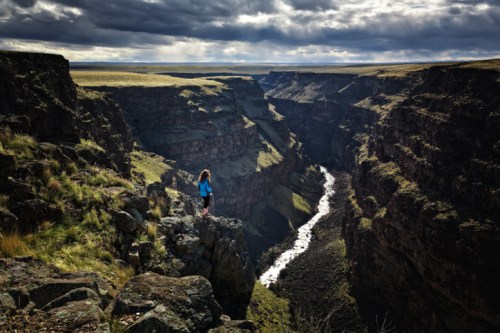 A person stands on a rocky ledge overlooking a deep canyon with a winding river below, under a dramatic sky.
