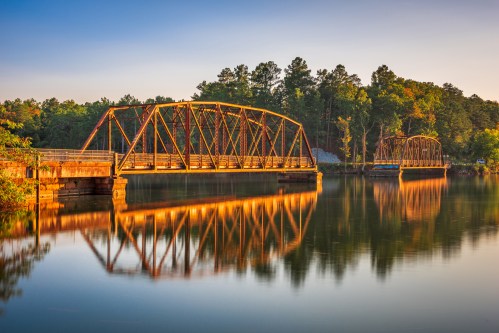 A rustic metal bridge spans a calm river, reflecting the surrounding trees and warm sunlight in the water.