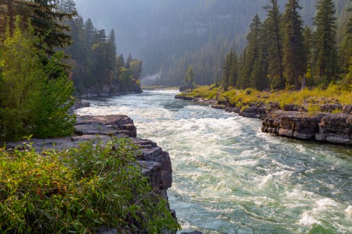 A winding river flows through a lush, forested landscape, surrounded by rocky cliffs and misty mountains.