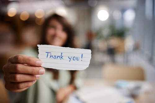 A person holds a handwritten note that says "Thank you!" in a blurred indoor setting.
