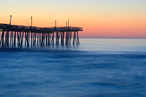 A serene pier extends over calm waters at sunset, with soft waves and a gradient sky of orange and blue hues.