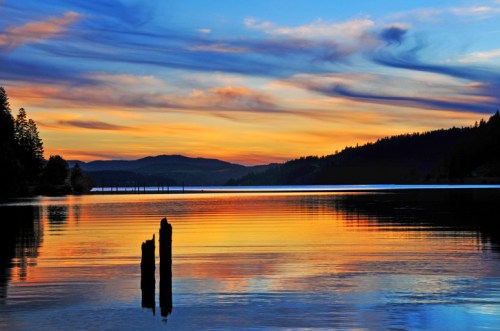 A serene sunset over a calm lake, with colorful clouds reflecting on the water and silhouettes of trees in the background.
