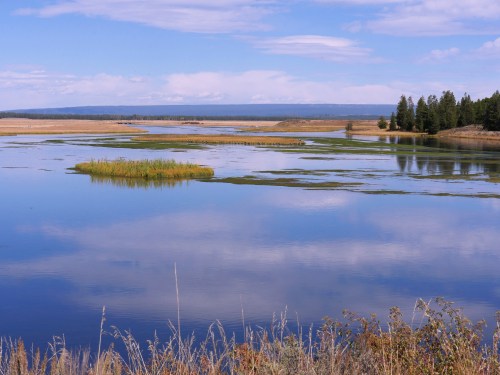 A serene landscape featuring a calm lake, grassy islands, and blue skies with scattered clouds.