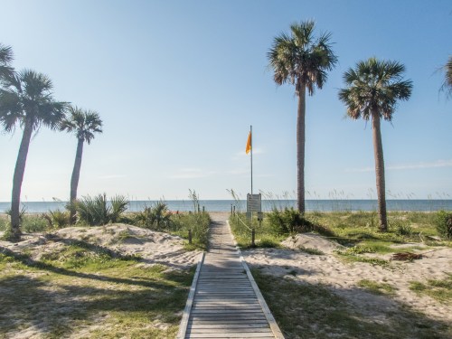 A wooden walkway leads to a beach, flanked by palm trees and a yellow flag, under a clear blue sky.