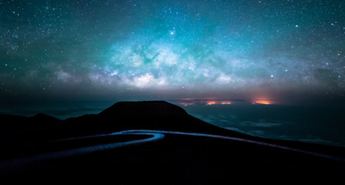 A winding road under a starry sky with the Milky Way and distant lights on the horizon.