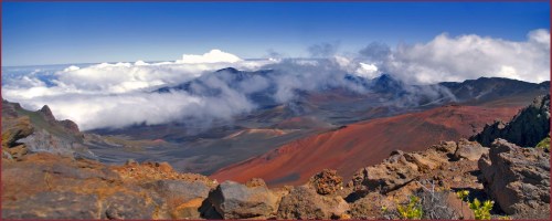 A panoramic view of a volcanic landscape with red earth, rocky formations, and clouds in a blue sky.
