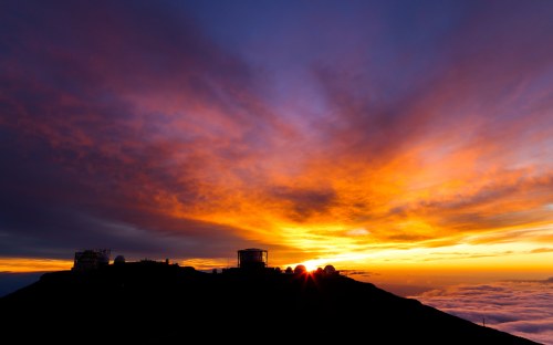 A vibrant sunset over a mountain silhouette, with clouds below and a building in the foreground.