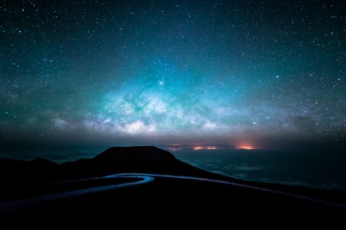 A stunning night sky filled with stars and the Milky Way, above a dark landscape with distant lights on the horizon.