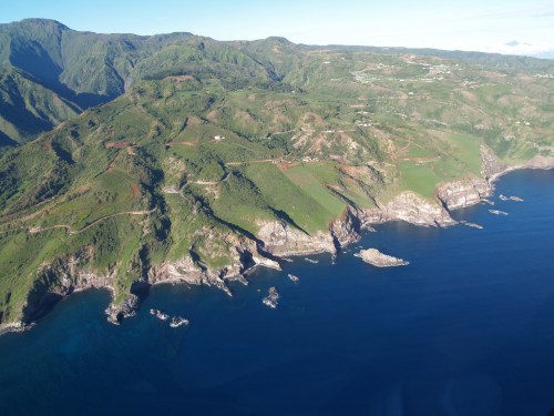 Aerial view of lush green hills meeting the blue ocean, with rocky cliffs and a clear sky in the background.