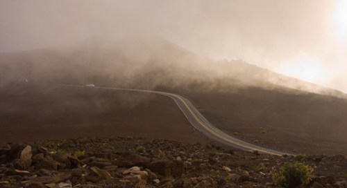 A winding road disappears into foggy, mountainous terrain, with rocky ground and soft light illuminating the scene.