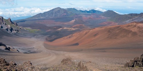 A vast volcanic landscape with colorful slopes, rocky terrain, and distant mountains under a clear blue sky.