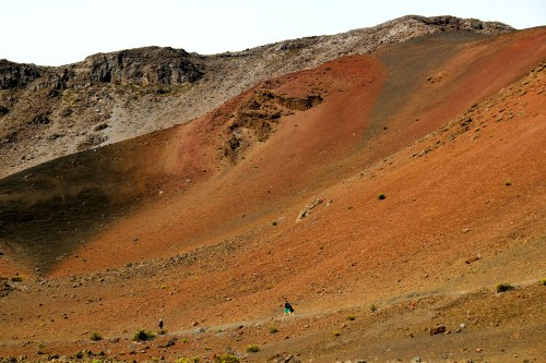 A hiker traverses a colorful, rugged mountain slope with red and brown hues under a clear sky.