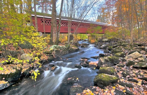 A red covered bridge spans a stream surrounded by autumn foliage and rocky banks.