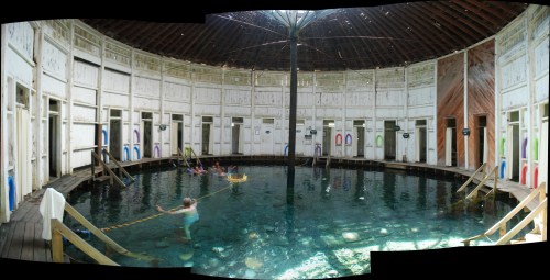 A panoramic view of a circular indoor pool with wooden walls, featuring people swimming and colorful doors around the perimeter.
