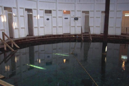 Interior of a circular room with a reflective water pool, wooden walkways, and doors along the walls.