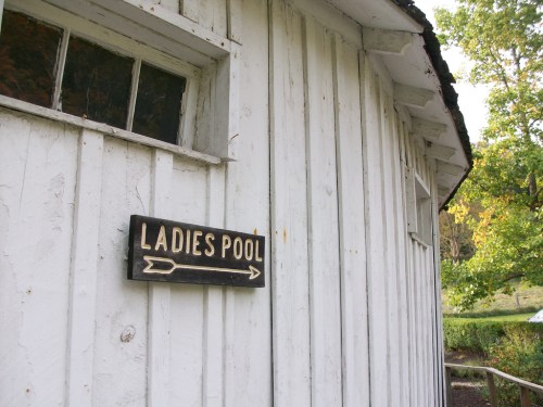 Sign for "Ladies Pool" on a white wooden building, with an arrow pointing to the right. Green trees in the background.
