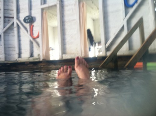 Feet submerged in water, with a rustic wooden structure and curtains in the background.
