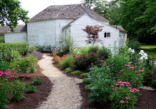 A gravel path winds through a colorful garden leading to a white house surrounded by lush greenery.