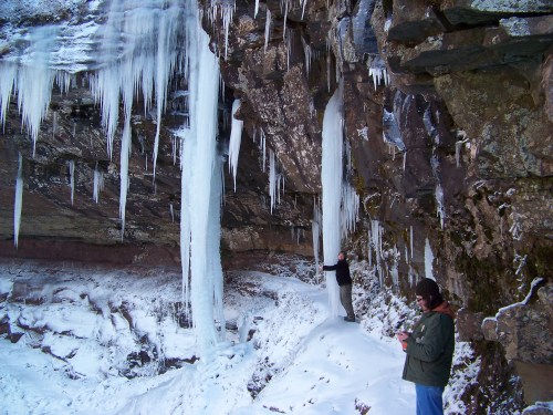 Two people stand near large icicles hanging from a rocky cliff, surrounded by snow-covered ground.