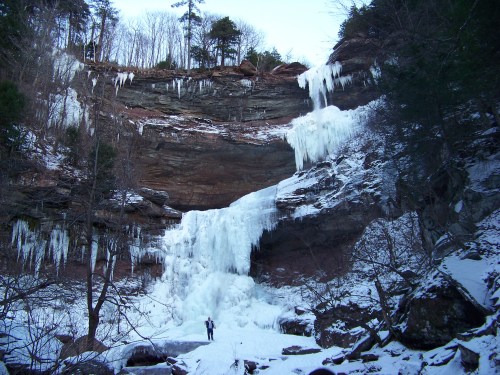 A frozen waterfall surrounded by icy cliffs and trees, with a person standing at the base for scale.
