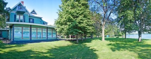 A large, green house with a wraparound porch surrounded by trees and a grassy area near a lake on a sunny day.