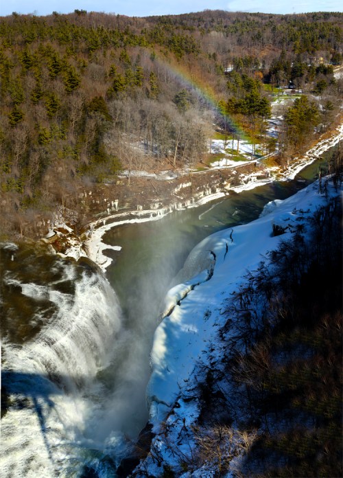A waterfall cascades over snowy cliffs, with a rainbow arcing above a forested landscape in the background.