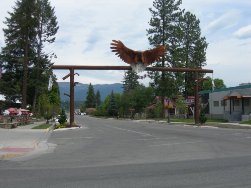 A large wooden arch with an eagle sculpture spans a quiet street lined with trees and buildings in a small town.