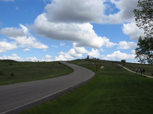 A winding road leads to a hilltop with people silhouetted against a cloudy sky and green grass.