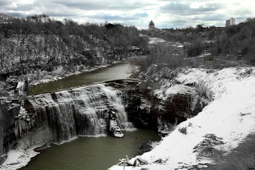 A frozen waterfall cascades into a river, surrounded by snow-covered trees and a cloudy sky.