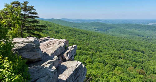 A panoramic view of lush green mountains and valleys from a rocky cliff under a clear blue sky.
