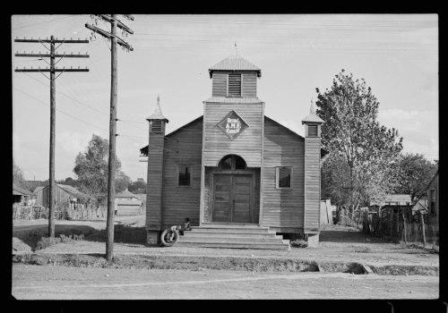 A historic wooden church with a triangular sign, surrounded by trees and power lines, in a rural setting.