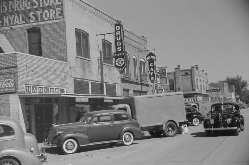 Historic street scene featuring vintage cars parked outside a drugstore and café in Harlem. Black-and-white photo.